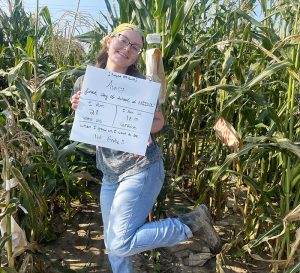 Avery Rayle holding a sign and posing for a picture in front of a field of corn.