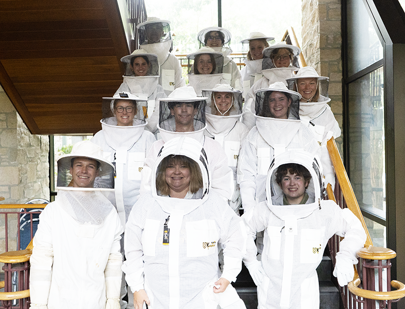 A group of individuals, dressed in beekeeping gear, smile at the camera for a picture.