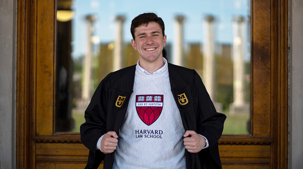 A student posing for a graduation photo in front of a large window.