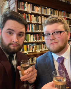 Two individuals smiling for the camera while standing in front of a bookshelf full of books.