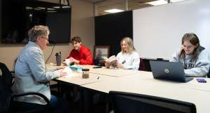 Students smiling while look at books and laptops while talking with their professor.