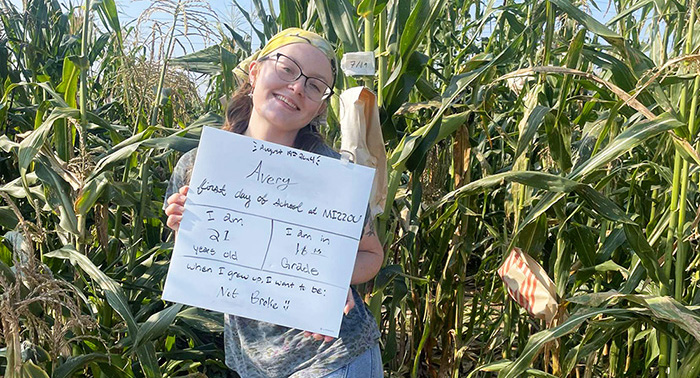 Avery Rayle holding a sign and posing for a picture in front of a field of corn.