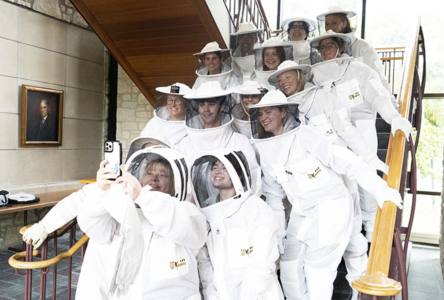 Students gather around their professor for a selfie. The students are wearing beekeeping safety gear.