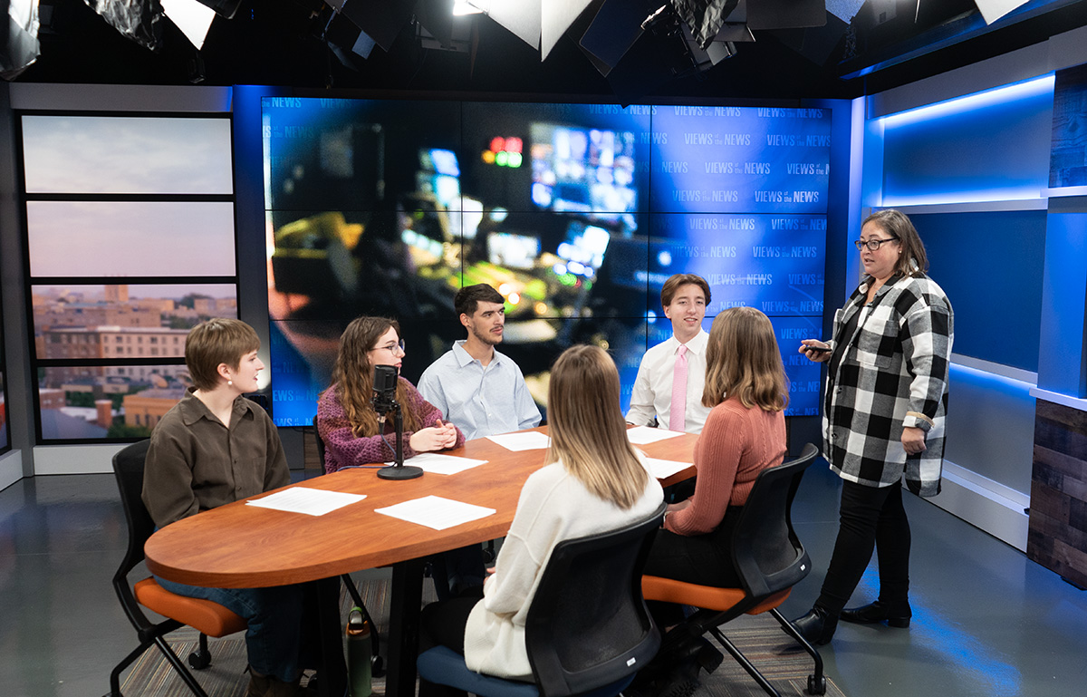 Students talking to their professors while gathered around a news desk.