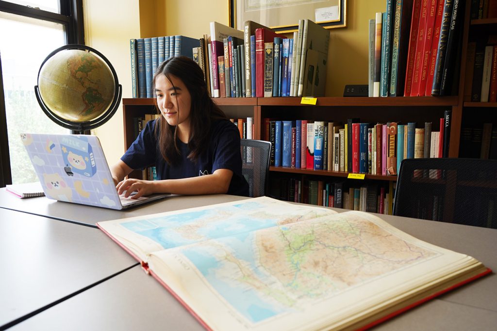 A student working on their laptop while in a library, with a large book opened beside her.