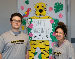 Two individuals pose for a picture in front of a sign that reads 