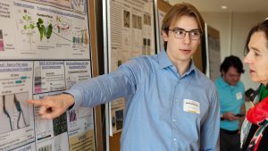 A student pointing at a research poster while talking to another individual.