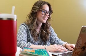Jane Ray smiling while working on her laptop in a small office.
