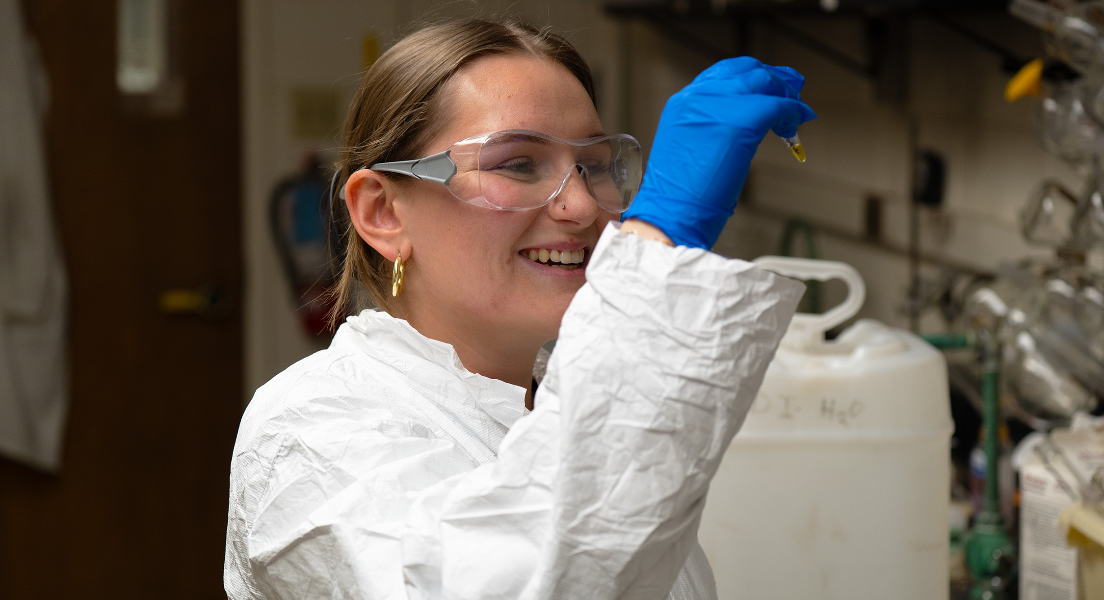 A student, wearing a lab coat, holding up a vial of liquid and staring at it.