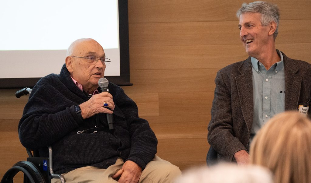 Bill Bondeson holding a microphone and talking while other smile and listen.