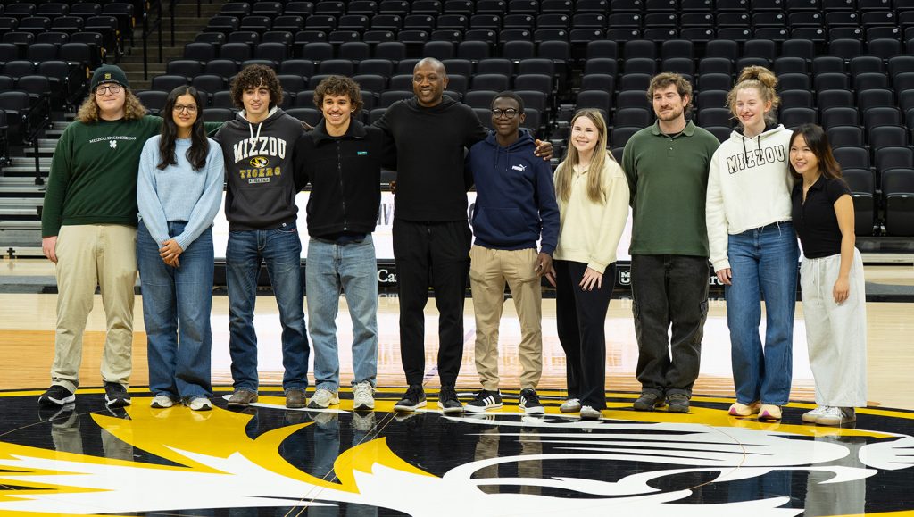 A group of students smiling while posing for a picture with Dennis Gates. They are all standing on the Mizzou basketball court.