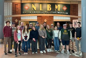A large group of students and a faculty member smiling while posing for a photo in front of the Negro League Baseball Museum.