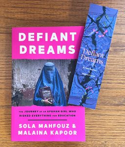 The book 'Defiant Dreams' sitting on a desk with a bookmark laying on top of it. The bookmark also has the name of the book -- 'Defiant Dreams' -- on it.
