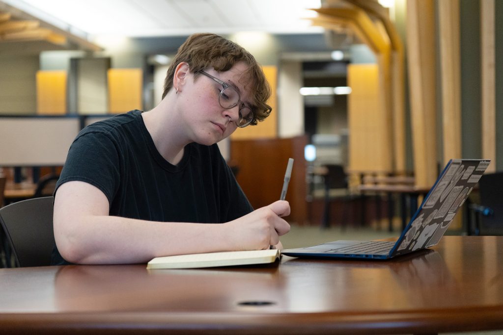 Alex Tyrrell writing in a notebook while sitting in the library.