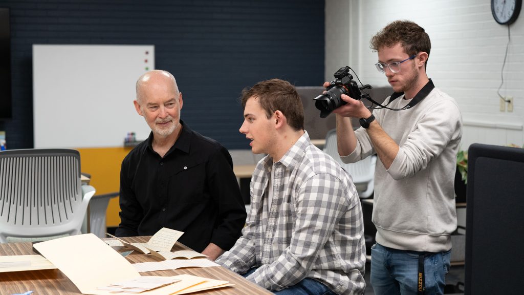 A faculty member talking with a student while another student films them.