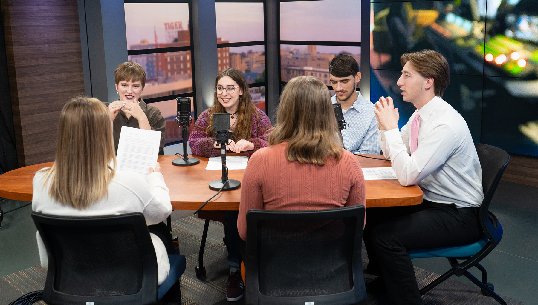 A group of students sitting at a desk and talking while preparing for a newscast.