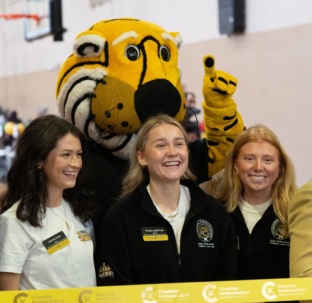 Three students smiling and laughing while Truman the Tiger stands behind them.