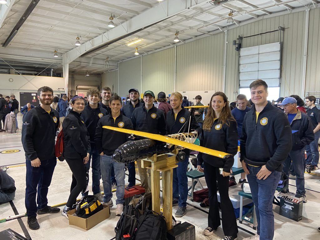 Group of students and faculty stand with a 3-D printed aeroplane in black and gold 