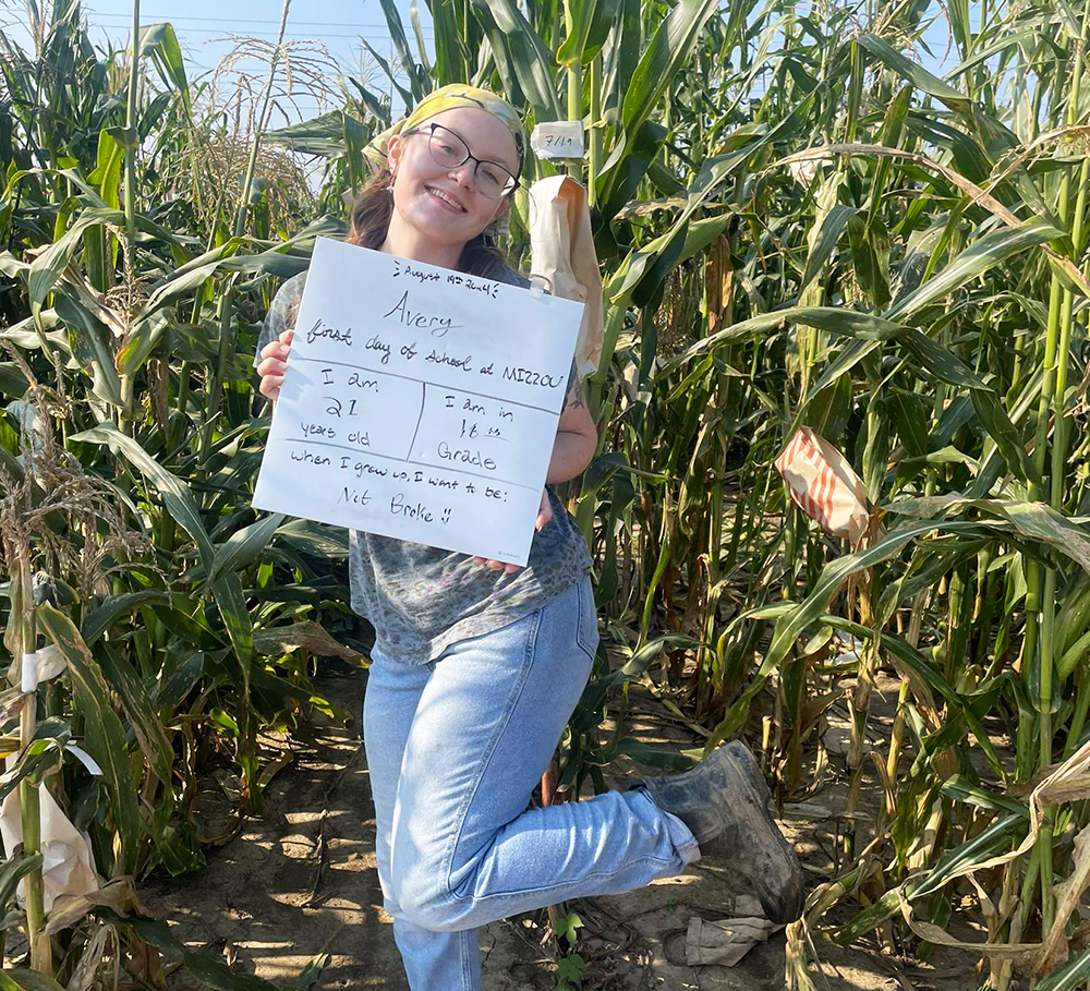 Avery Rayle holding a research poster while posing for a picture in front of a field of corn.