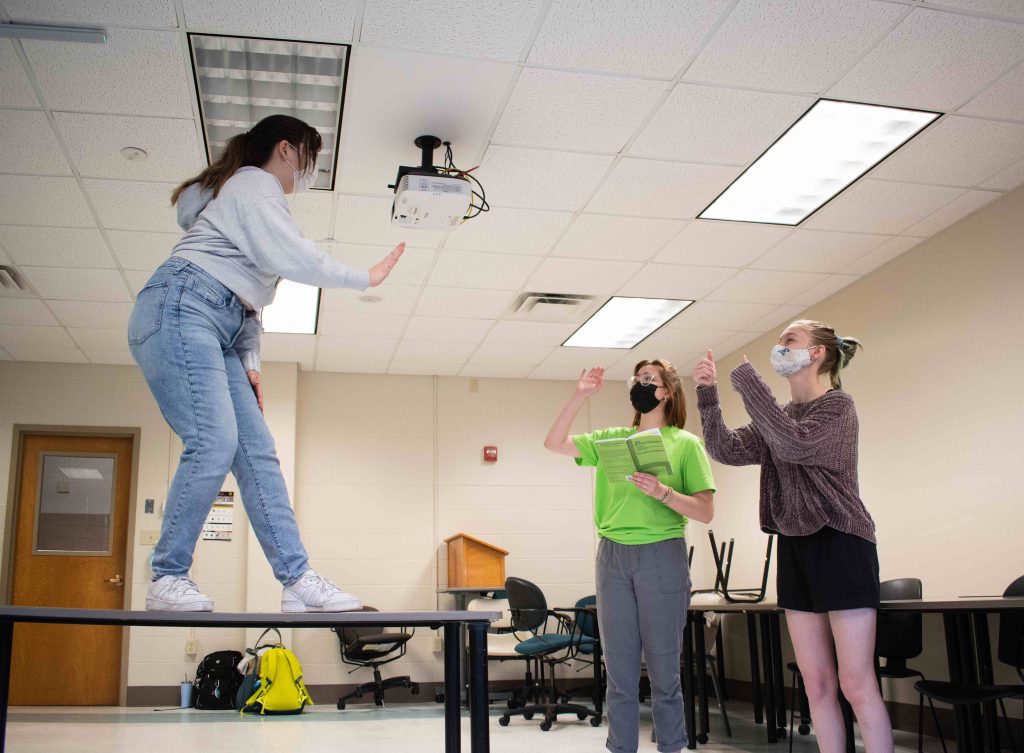 One Student stands on a table acting while two other students stand on the floor in response
